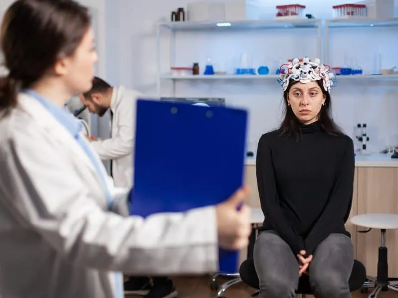 Medic researcher pointing at clipboard explaning treatment against brain disease to patient with eeg headset. Woman sitting in neurological scientific laboratory treating nervous system dysfunctions.