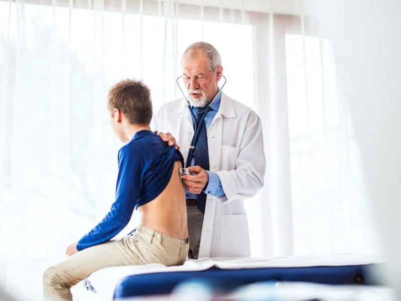 Senior male doctor examining a small boy with stethoscope in his office.