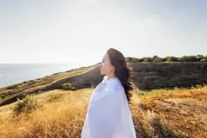 A young handsome Asian woman wrapped in a white sheet and looks at the seascape. A charming Korean woman enjoys nature. Profile of an attractive girl in the rays of the sun at sunset outdoors.
