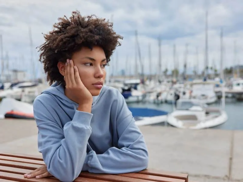 Beautiful young African American woman admires yachts and calm sea water spends free time on harbor pier dressed in hoodie dreams about summer journey on vacation walks outdor after quarantine