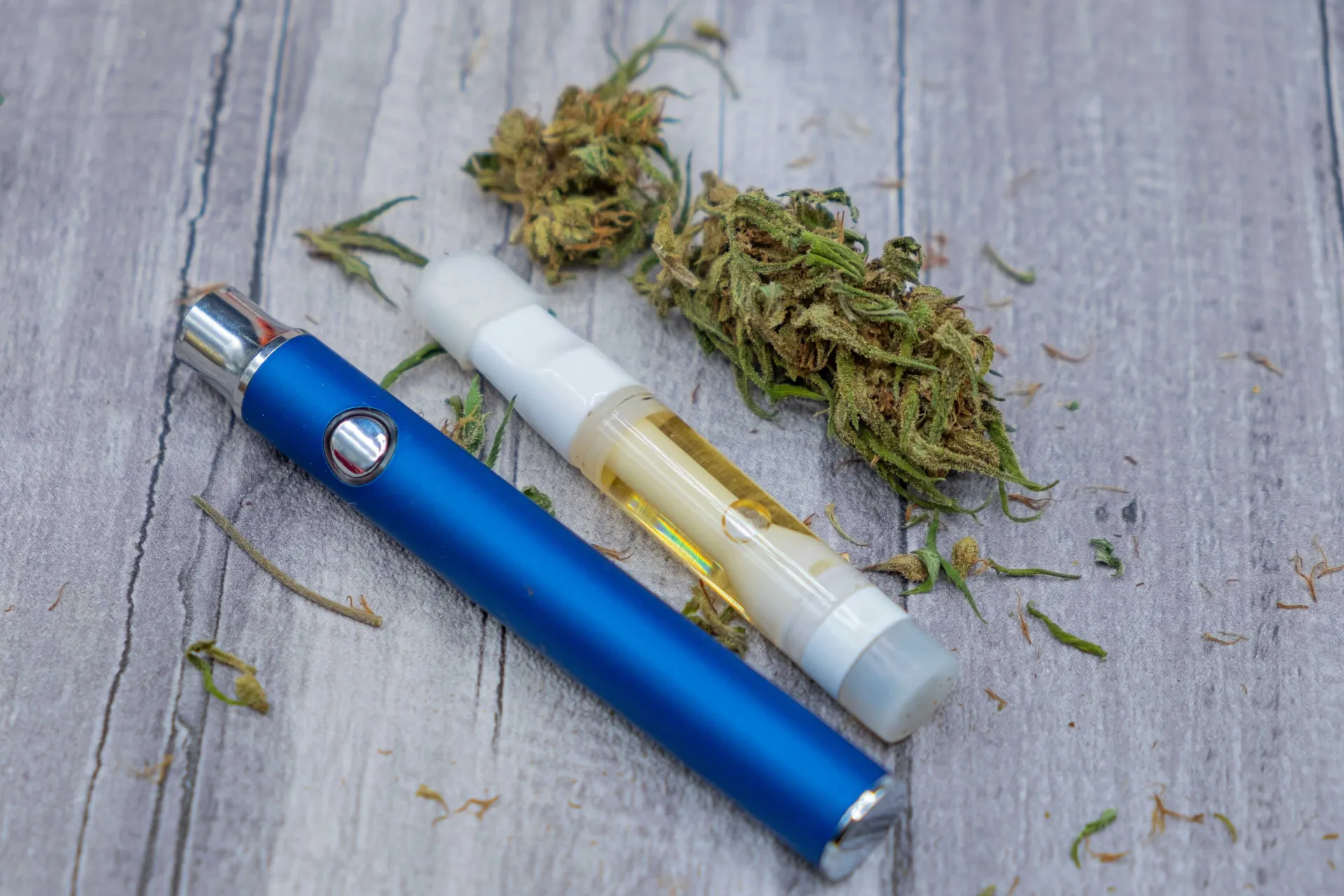 A blue vape pen and a white cartridge filled with cannabis oil, alongside dried cannabis buds on a wooden surface.