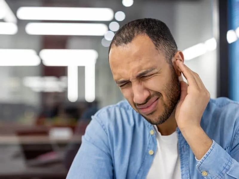 Close-up photo of a young Latin American man in the office, grimacing in pain, holding his ear with his hand.