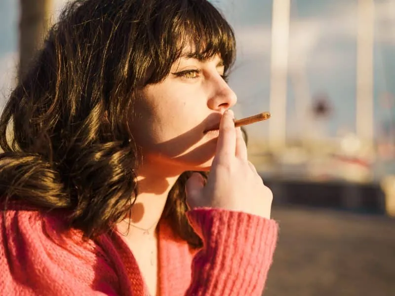 Young woman smoking by the sea - A young woman smokes by the sea, reflecting on youth's vices. The calm setting contrasts with her intense gaze, highlighting the complexities of modern indulgences.