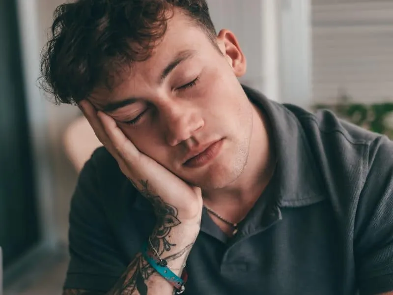 Handsome young caucasian man resting with closed eyes and head on his hand, portraying a sense of fatigue or calm reflection. He has visible tattoos and a casual outfit,