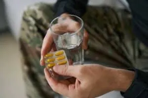 A person holding a blister pack of yellow capsules in one hand and a glass of water in the other