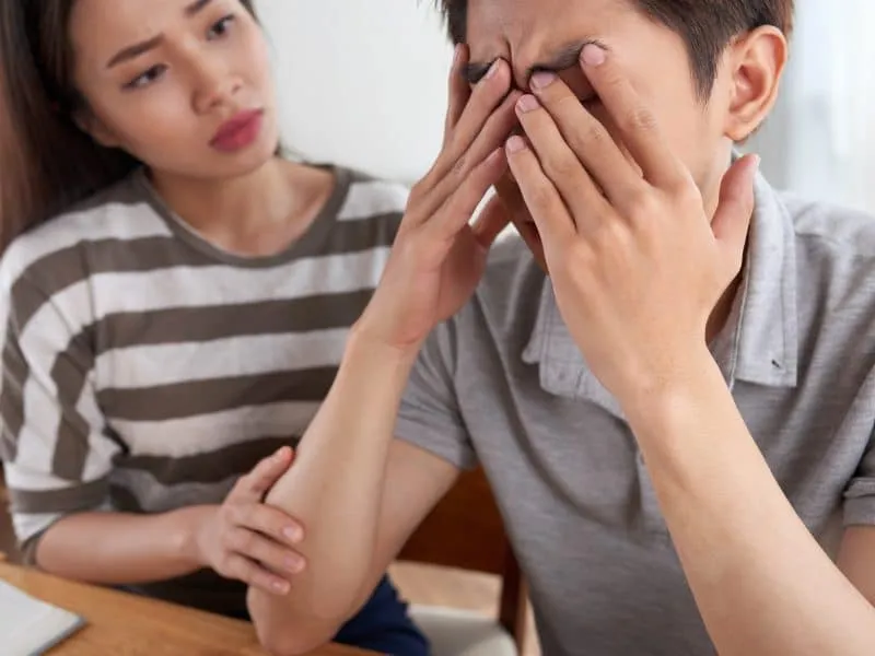 Unrecognizable sad man covering his face with hands while his pretty Asian girlfriend trying to cheer him up