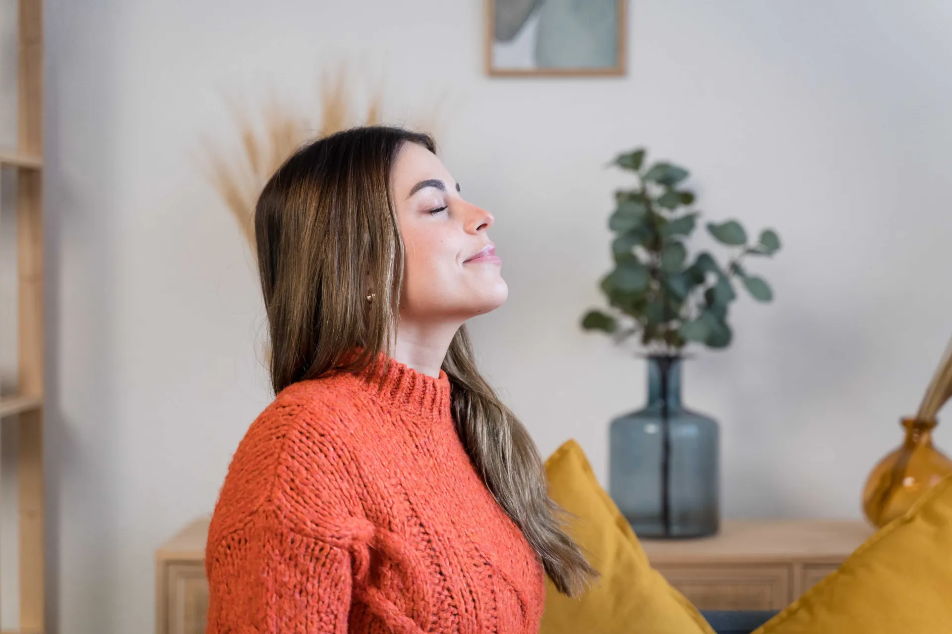A woman in a cozy orange sweater sits against a bright, neutral backdrop, with a vase of greenery and decorative pillows nearby.