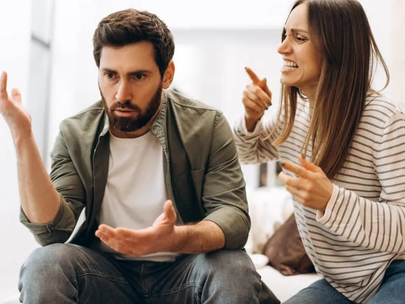 Young man and woman sitting on a sofa at home, engaged in a heated argument, gesturing expressively and displaying frustration over their relationship issues. Emotions running high