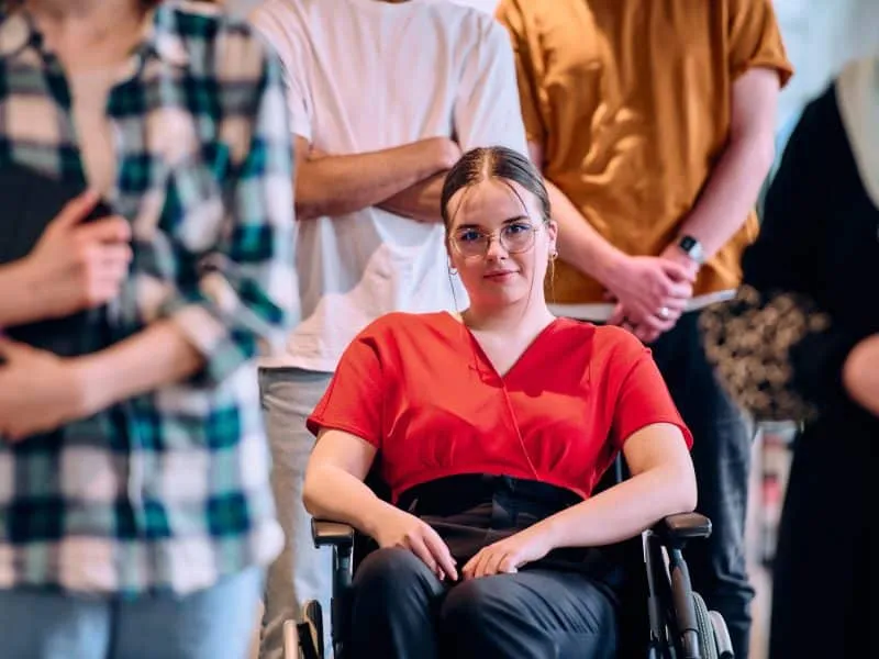 A diverse group of young business people walking a corridor in the glass-enclosed office of a modern startup, including a person in a wheelchair and a woman wearing a hijab.