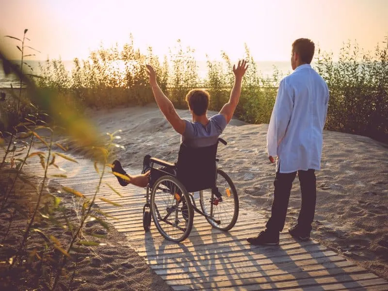 A man on wheelchair and his nurse enjoying sunrise on a beach.