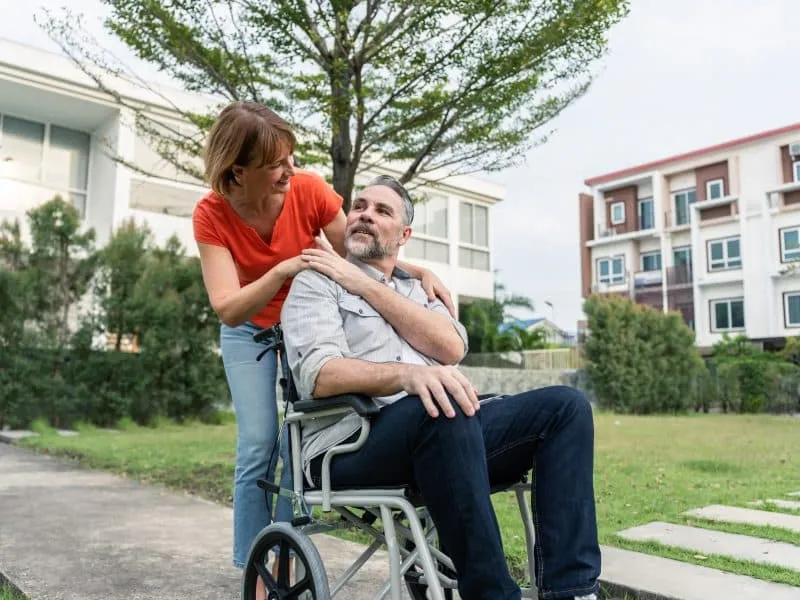 Caucasian senior woman support husband on wheelchair outdoor in garden. Attractive elderly mature wife take care and walking with older man patient, doing physical therapy for health togethet in park.