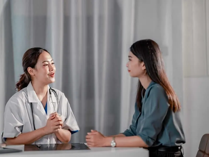 A healthcare professional in a white coat discusses with a patient at a clinic, with medical equipment in the background.