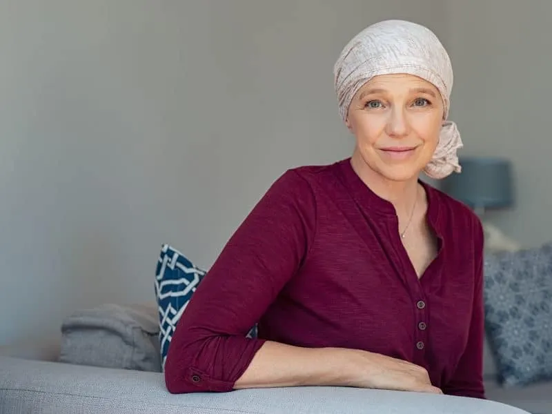 Mature woman with cancer in pink headscarf smiling sitting on couch at home. Smiling woman suffering from cancer sitting after taking chemotherapy sessions. Portrait of mature lady facing side-effects of hair loss, copy space.