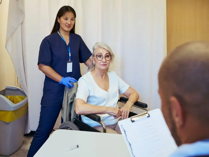 Doctor and nurse assisting elderly patient in wheelchair during medical consultation in hospital room, demonstrating teamwork and healthcare
