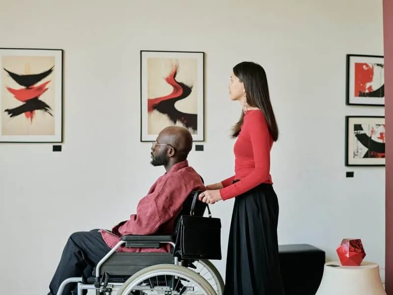 Man sitting in wheelchair and woman standing next to him are observing abstract artworks in a modern gallery space with clean design