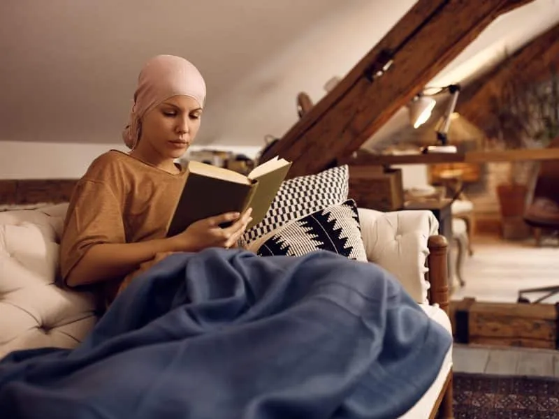 Young woman with cancer reading a novel and wearing headscarf while relaxing on the sofa.