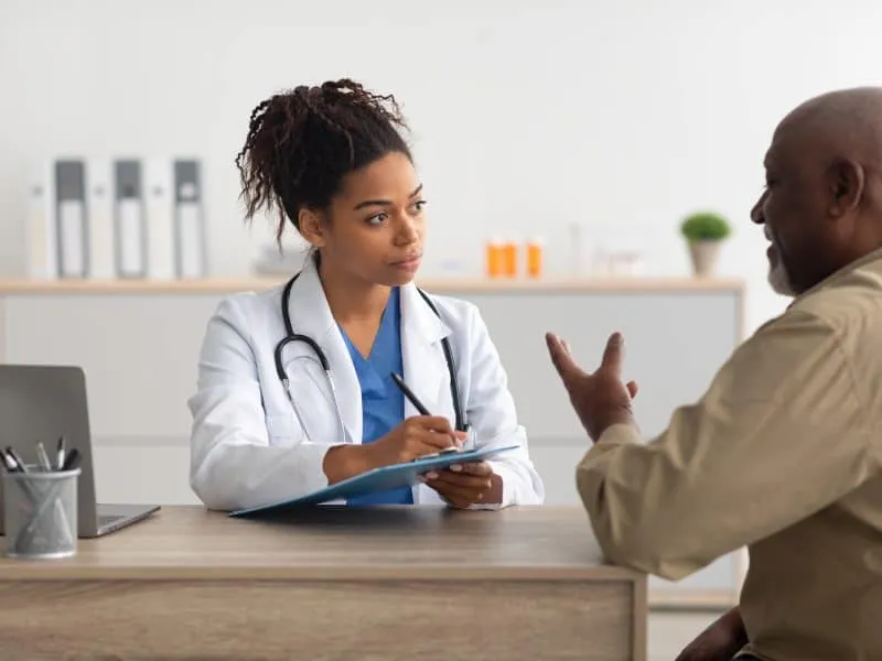 General Practitioner Appointment. Portrait of focused black female doctor listening to talking patient, discussing diagnosis and treatment, writing. Specialist giving consultation to elderly man