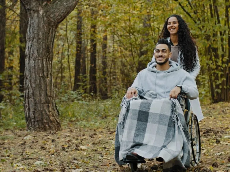 Smiling nurse girl in medical gown pushing young guy in wheelchair walking in hospital park enjoy walk in fresh air communicate feel happiness woman taking care disabled patient during rehabilitation. High quality 4k footage