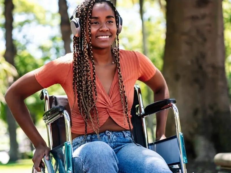 African American woman in a wheelchair smiling and listening to music with headphones at the park.