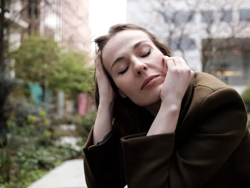 Portrait of relaxed woman with eyes closed sitting on table outdoors. She is between buildings and trees wearing a coat.