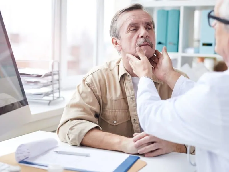 Over shoulder view of senior doctor sitting at table and touching neck of mature man while doing neck lump examination