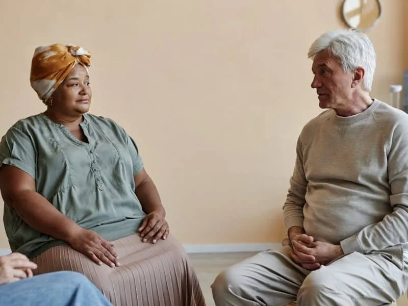 Group of senior people sitting in circle at mental health support group with focus on white haired man sharing