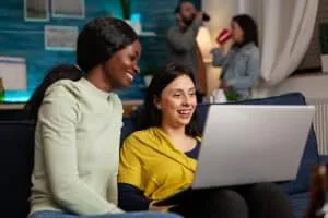Multi-ethnic friends socializing while watching online funny videos on laptop resting on sofa. In background two womens drinking beer enjoying time spend together during entertainment party.