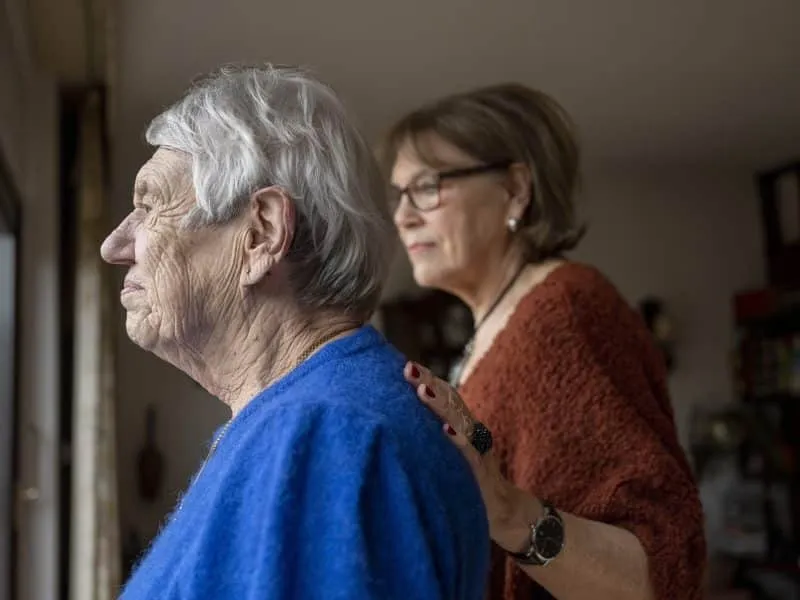Two elderly friends standing looking out of the window consoling one another