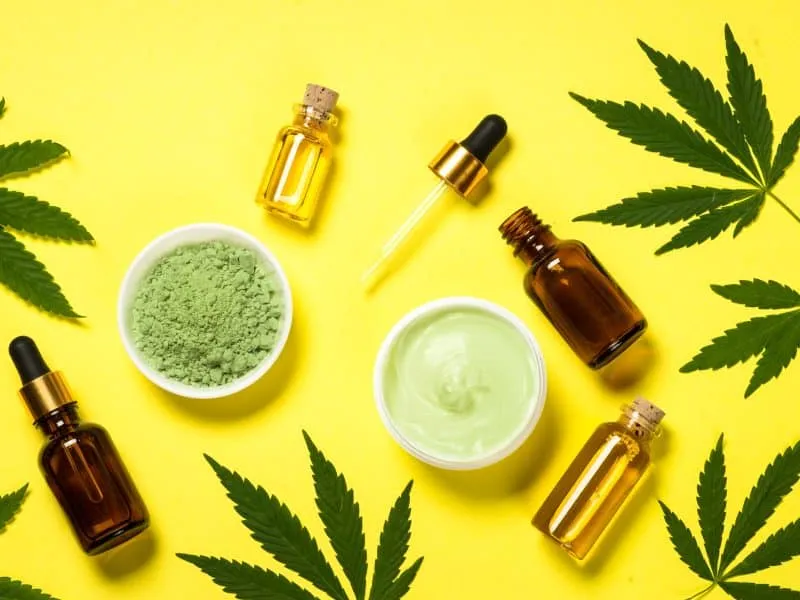 Cannabis product for health. Cannabis oil, cream, pills and powder with fresh cannabis leaves at yellow table. Top view.