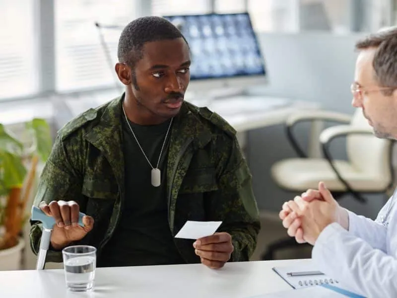 Mature Caucasian doctor giving prescription and explaining something to young Black military man with injury using stick to walk