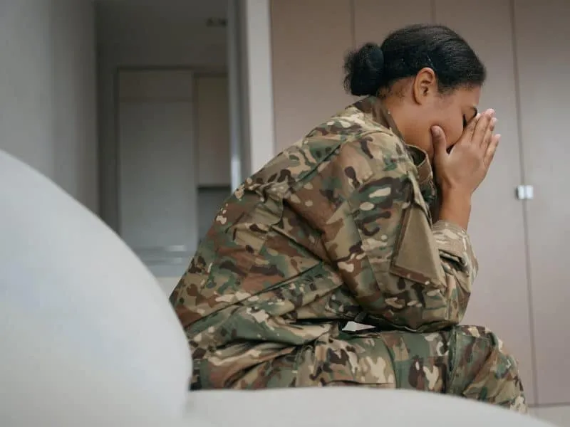 Tired african american female soldier sits alone in a bright room, she is wearing camouflage clothes