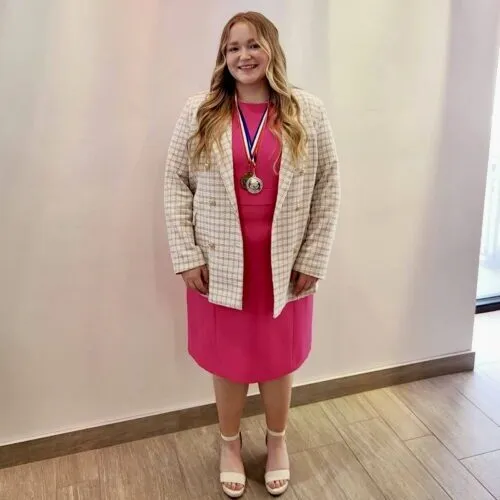 Frink.Presley_Gold Young woman stands in front of a multicolored background, smiling